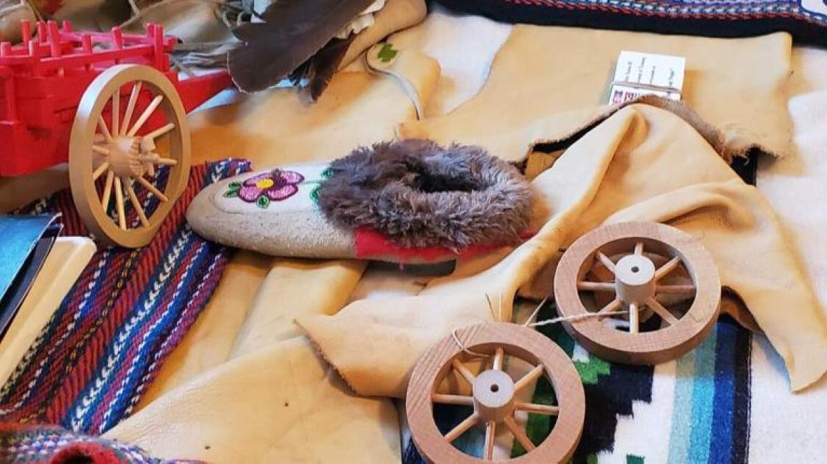 On a table are metis weaving tools, a loom, a woven sash, beaded and fur-trimmed moccasins, rawhide and feathers