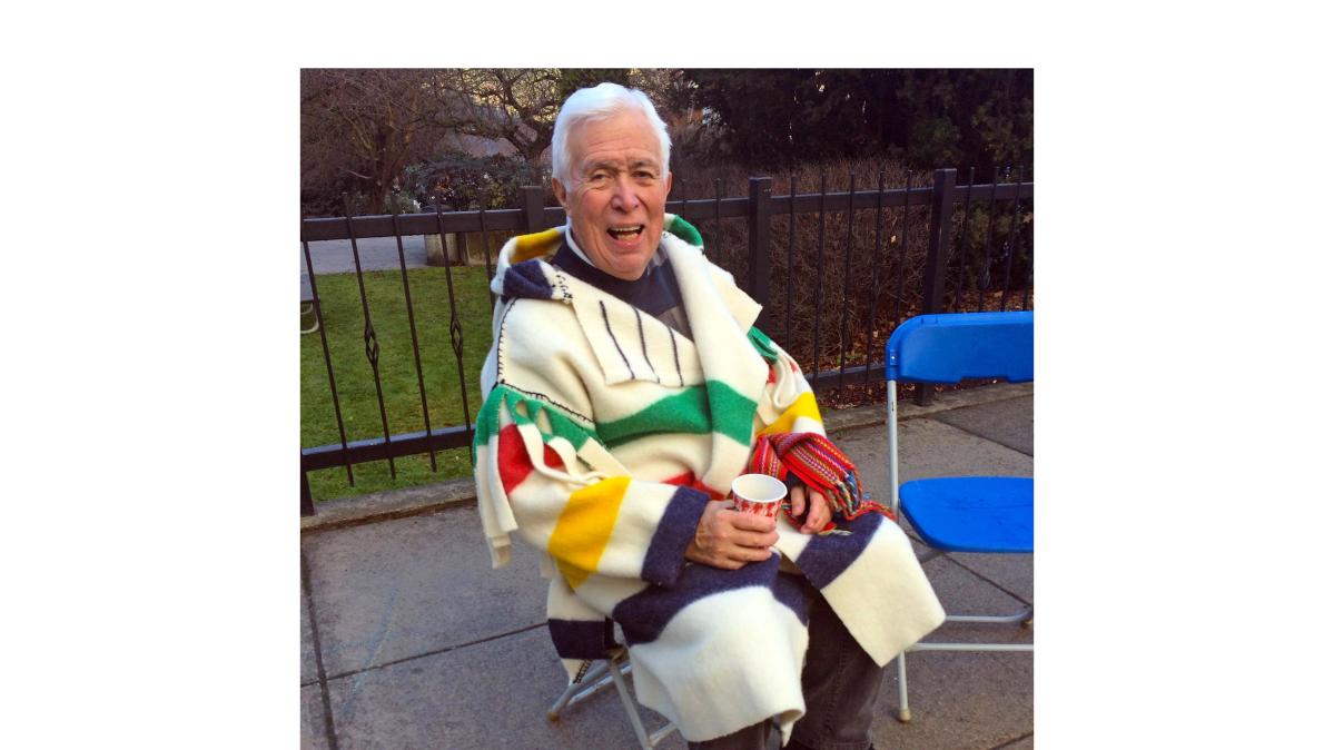 An elder in a blanket coat sits on a chair in a courtyard.