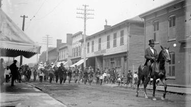 A black and white historic photo of a man in a top hat riding a horse at the front of a parade of people