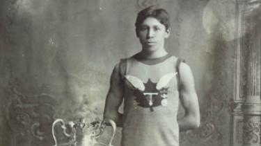 A black and white photograph of a young man in a singlet and white shorts standing in front of a large trophy.