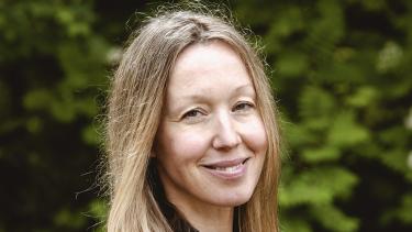 A woman with dark blonde long hair wearing a black shirt smiles in front of a green bush outdoors.