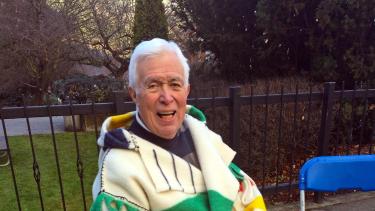 An elder wearing a Hudson's Bay blanket jacket sits in a folding chair in an outdoor courtyard.