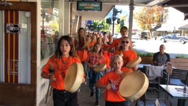 Children in orange shirts walk down a town sidewalk drumming