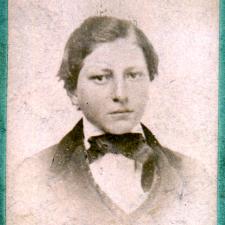 A faded photograph of a teen with short brown hair wearing a high collar, bow-tie and suit jacket 