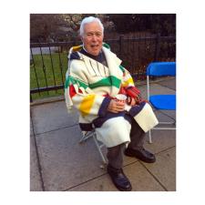 An elder in a blanket coat sits on a chair in a courtyard.